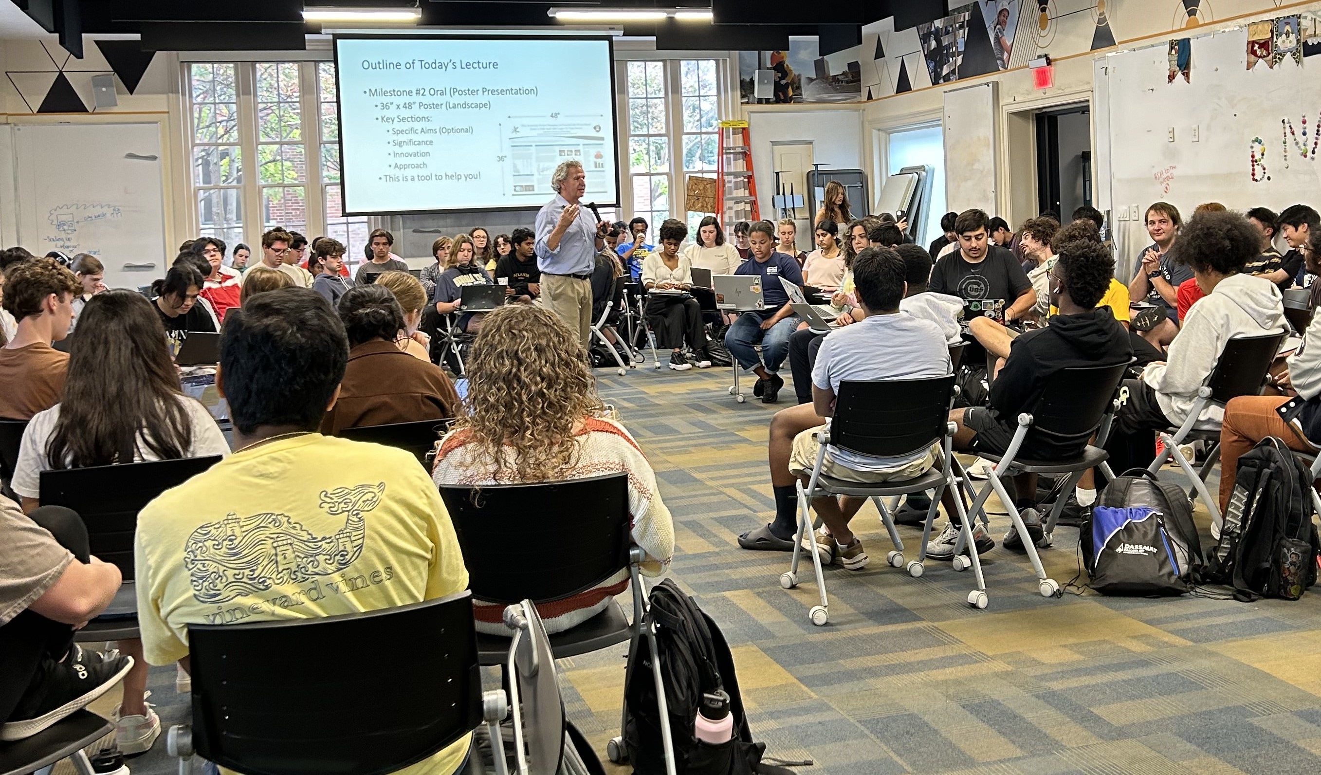 Rudy Gleason, an older white man with gray hair, stands in the middle of students holding a microphone and gesturing as he speaks to students