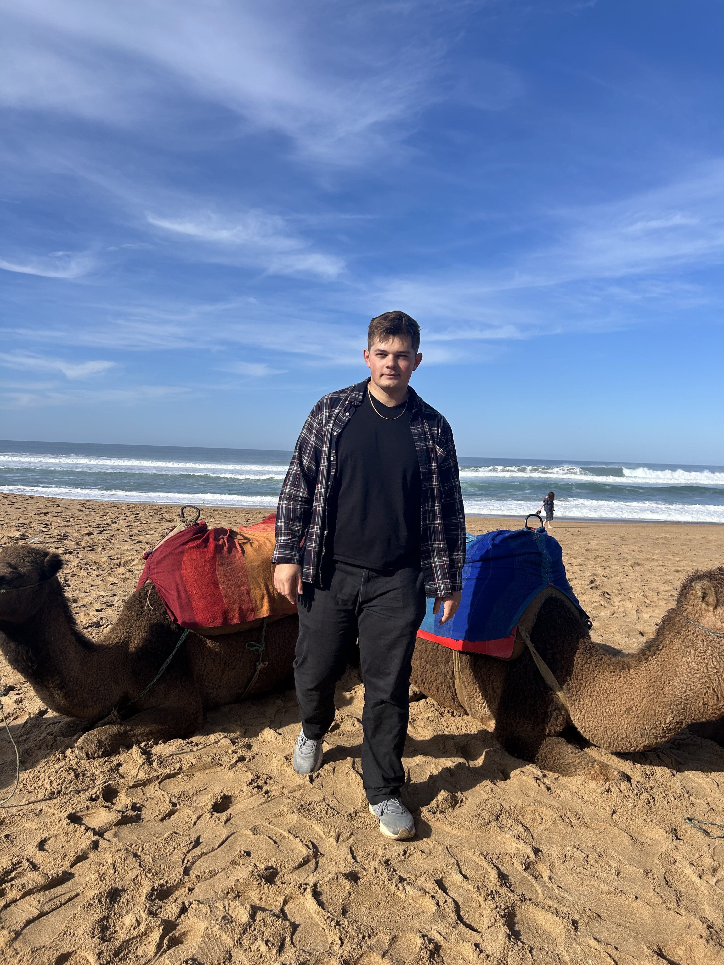 Young white man with brown hair wearing dark clothing standing in front of two camels who are laying down with an ocean in the background